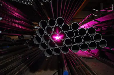 A stack of metal pipes, with pink light shining through the center, is arranged and stored in an industrial setting.